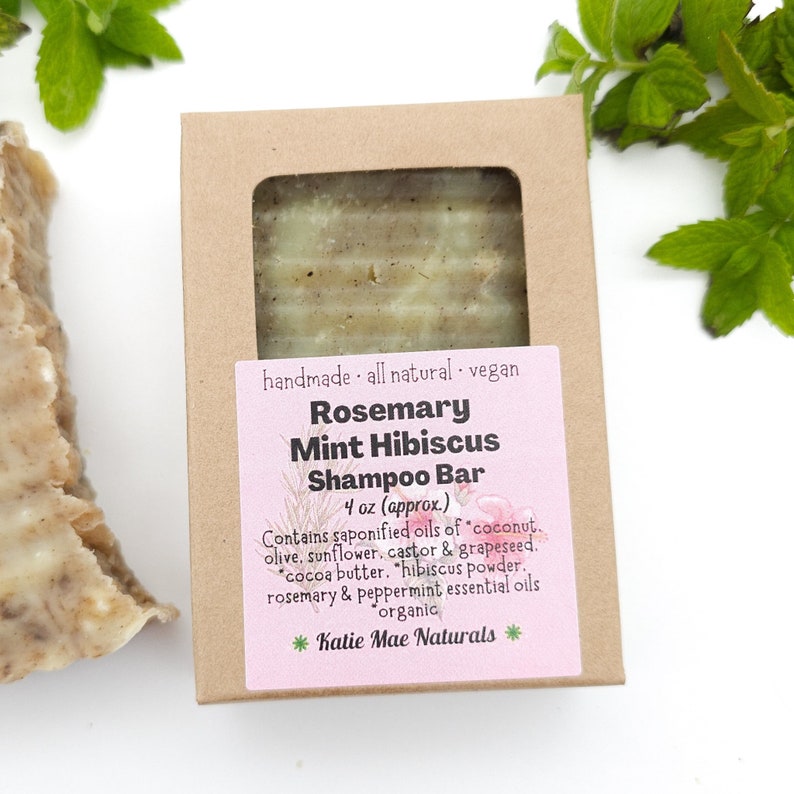 Może przedstawiać: Brązowe pudełko kartonowe z r&oacute;żową etykietą, na kt&oacute;rej widnieje napis "Rosemary Mint Hibiscus Shampoo Bar" oraz lista składnik&oacute;w. Etykieta zawiera r&oacute;wnież nazwę marki "Katie Mae Naturals".