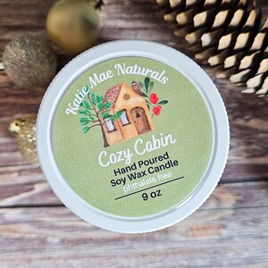 Puede incluir: Un tarro de vela blanco con una etiqueta que dice "Katie Mae Naturals Cozy Cabin". La etiqueta presenta un dibujo de una caba&ntilde;a, un &aacute;rbol y bayas. La vela es de cera de soja vertida a mano, sin ftalatos, y pesa 255 gramos.