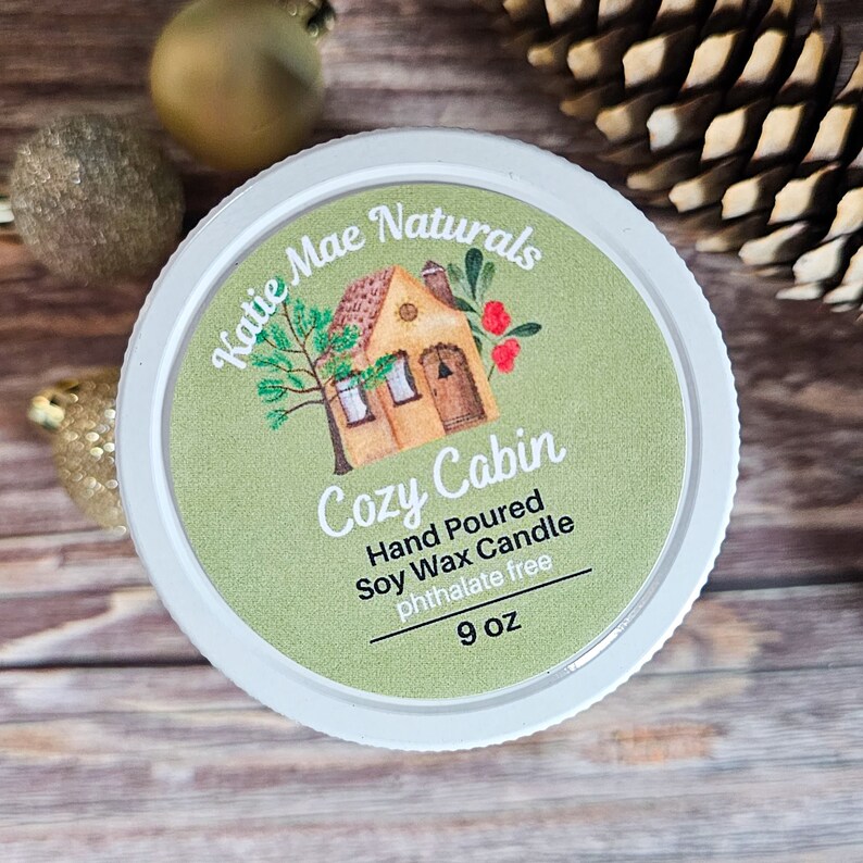 Puede incluir: Un tarro de vela blanco con una etiqueta que dice "Katie Mae Naturals Cozy Cabin". La etiqueta presenta un dibujo de una caba&ntilde;a, un &aacute;rbol y bayas. La vela es de cera de soja vertida a mano, sin ftalatos, y pesa 255 gramos.