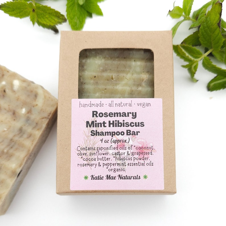 Może przedstawiać: Brązowe pudełko kartonowe z białą etykietą, na kt&oacute;rej widnieje napis: "Rosemary Mint Hibiscus Shampoo Bar 4 oz (approx.) Ręcznie robiony. Naturalny. Wegański. Zawiera mydlane oleje z kokosa, oliwy z oliwek, słonecznika, rycyny i pestek winogron. Masło kakaowe. Proszek z hibiskusa. Olejki eteryczne z rozmarynu i mięty pieprzowej. Organiczny. *Katie Mae Naturals*"