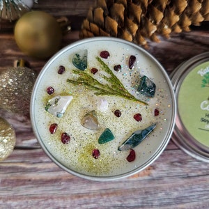 May include: A white soy candle with a green sprig of greenery and various colored crystals embedded in the wax. The candle is in a silver tin with a green label that reads "Handmade Soy Candle".