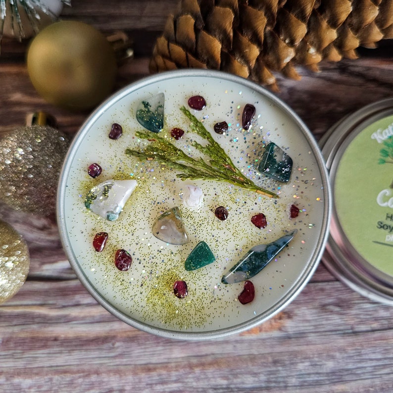 May include: A white soy candle with a green sprig of greenery and various colored crystals embedded in the wax. The candle is in a silver tin with a green label that reads "Handmade Soy Candle".