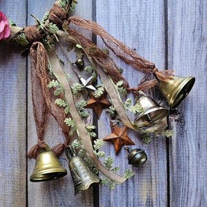 May include: A rustic wreath decorated with brown and green fabric, lace, and five antique silver bells. The wreath is adorned with five star-shaped embellishments.