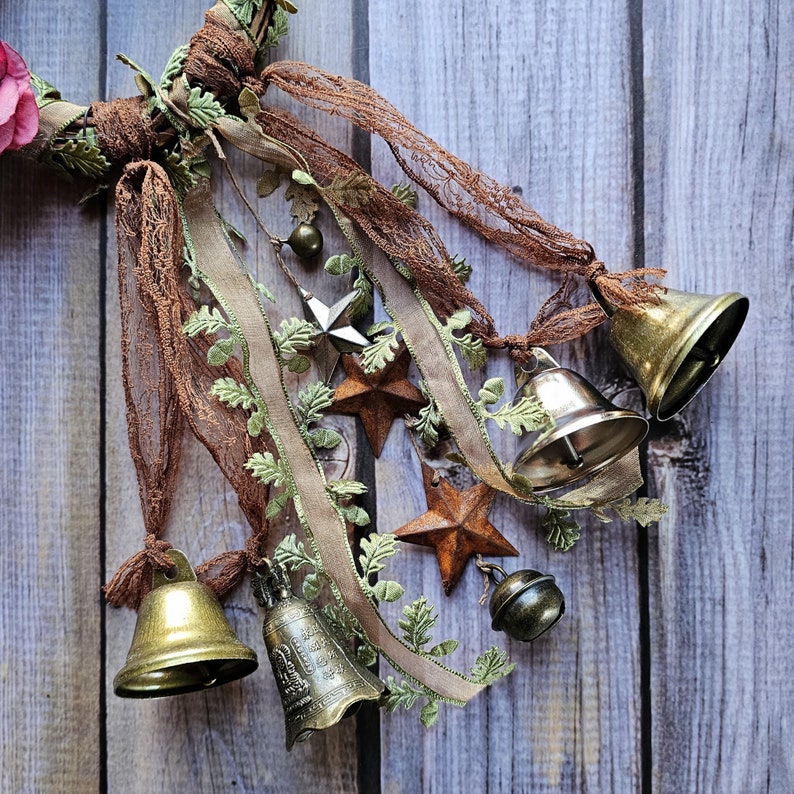 May include: A rustic wreath decorated with brown and green fabric, lace, and five antique silver bells. The wreath is adorned with five star-shaped embellishments.