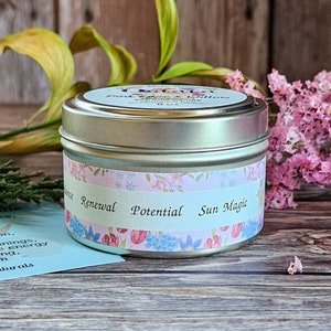 Peut inclure: Une bougie en bo&icirc;te argent&eacute;e avec un motif floral et les mots "Renouvellement", "Potentiel" et "Magie du soleil" sur l'&eacute;tiquette. La bougie est entour&eacute;e de fleurs roses et de feuillage vert.