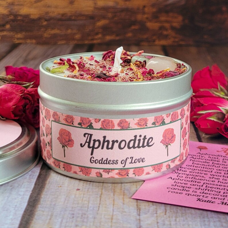 May include: A silver tin candle with a pink and white label that reads "Aphrodite Goddess of Love". The candle is decorated with rose petals and a clear crystal.