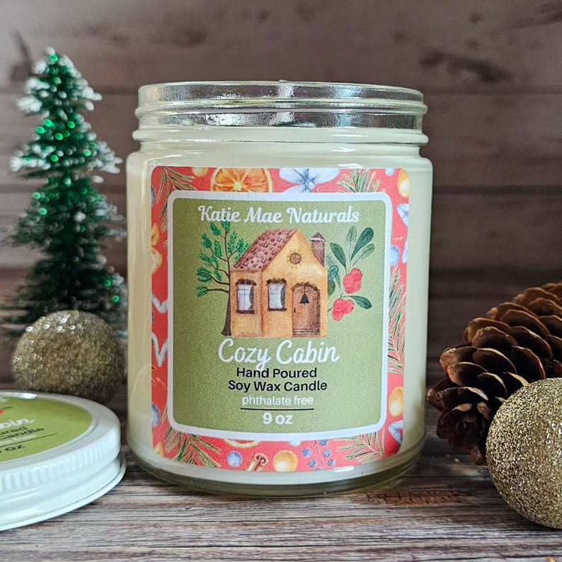 Puede incluir: Una vela en tarro de vidrio con una etiqueta que dice "Cozy Cabin" y "Katie Mae Naturals". La etiqueta presenta una ilustraci&oacute;n de caba&ntilde;a y est&aacute; rodeada de un dise&ntilde;o festivo. La vela es de 255 gramos.