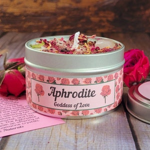 May include: A silver tin candle with a pink and white label that reads "Aphrodite Goddess of Love". The candle is decorated with dried rose petals and crystals.