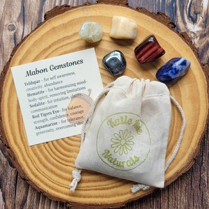 May include: A set of six gemstones in a small white drawstring bag with a green logo that says "Katie Mae Naturals". The gemstones are feldspar, hematite, sodalite, red tiger's eye, aquamarine, and a clear crystal. The bag is sitting on a wooden surface. Mabon Gemstones are listed with their properties on a card next to the bag.