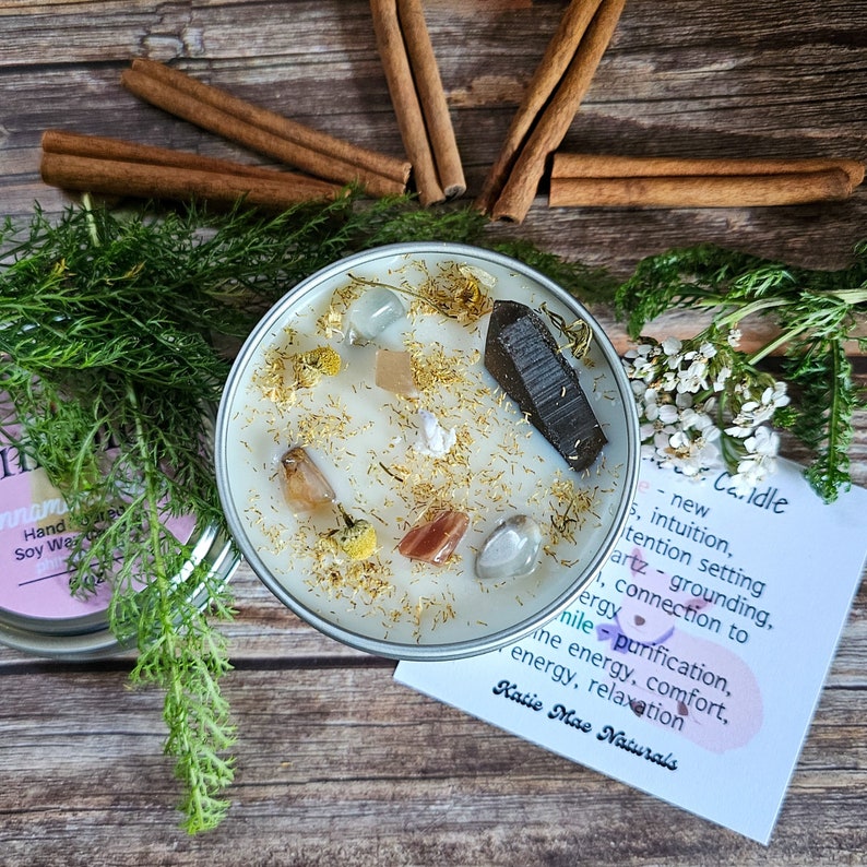May include: A white candle with gold glitter and various crystals embedded in the wax. The candle is in a clear glass jar with a silver lid. The candle is surrounded by cinnamon sticks and greenery. The text on the card reads "new intuition, intention setting, quartz - grounding, connection to energy, purification, the energy, comfort, energy, relaxation Katie Mae Naturals".