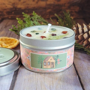May include: A small, round, silver tin candle with a green and white wax top. The candle has a label with a pink background and a cartoon illustration of a small house with a chimney. The text on the label reads "Cozy Cabin".