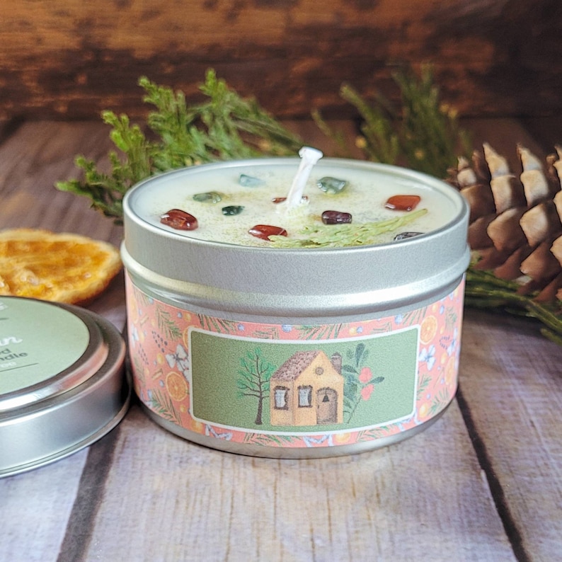 May include: A small, round, silver tin candle with a green and white wax top. The candle has a label with a pink background and a cartoon illustration of a small house with a chimney. The text on the label reads "Cozy Cabin".