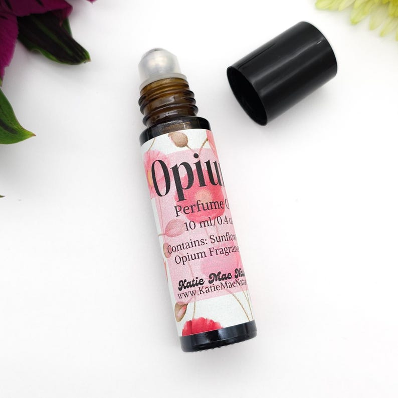 Puede incluir: Un peque&ntilde;o frasco de vidrio con un aplicador de bola negro. El frasco est&aacute; etiquetado como "Opium Perfume Oil" y contiene 10 ml / 0.4 fl oz. La etiqueta tambi&eacute;n incluye el texto "Contiene: Aceite de girasol Fragancia de opio" y "Katie Mae Naturals" con la direcci&oacute;n del sitio web "www.KatieMaeNaturals.com".