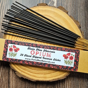 May include: A bundle of black incense sticks with gold tips, presented on a wooden slice. The packaging features the text "Katie Mae Naturals OPIUM 20 Hand Dipped Incense Sticks" with poppy illustrations and a decorative border.