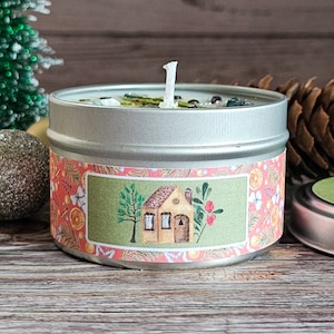 May include: A silver tin candle with a green and pink label. The label features a small illustrated house with a brown roof and a yellow door. The candle is surrounded by pine needles and a small Christmas tree.