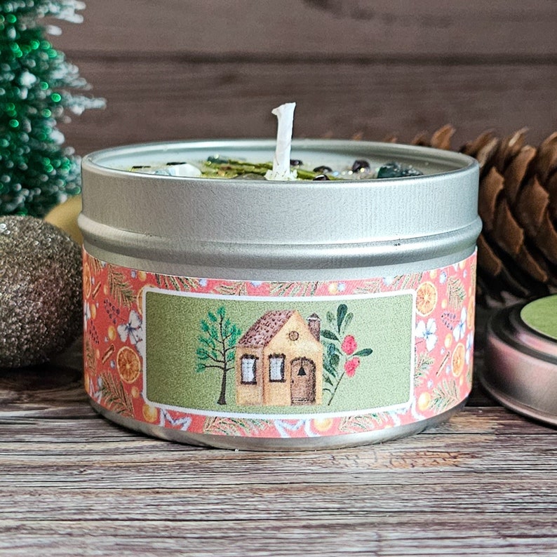 May include: A silver tin candle with a green and pink label. The label features a small illustrated house with a brown roof and a yellow door. The candle is surrounded by pine needles and a small Christmas tree.