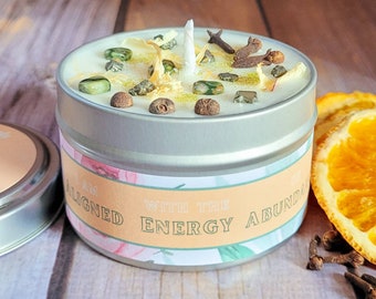 May include: A silver tin candle with a white wax top and a floral design label that reads "I am aligned with the energy abundance". The candle is surrounded by orange slices, cloves, and green and brown crystals.
