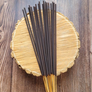 May include: A bundle of dark brown incense sticks with golden tips, arranged on a light brown, circular wooden slab. The background is a dark brown wooden surface. The incense sticks are tightly packed together, creating a visually appealing display.