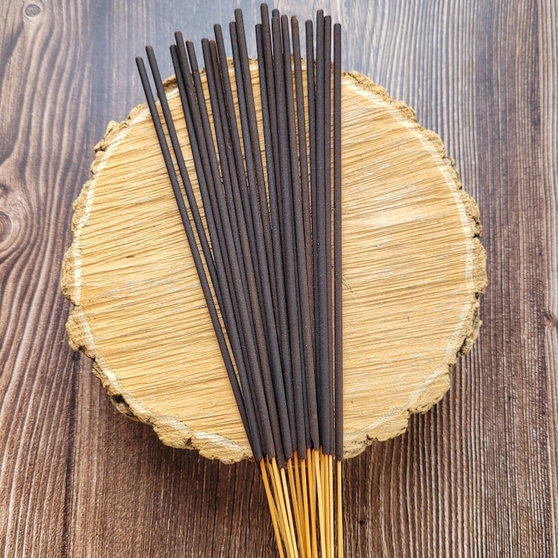 May include: A bundle of dark brown incense sticks with golden tips, arranged on a light brown, circular wooden slab. The background is a dark brown wooden surface. The incense sticks are tightly packed together, creating a visually appealing display.