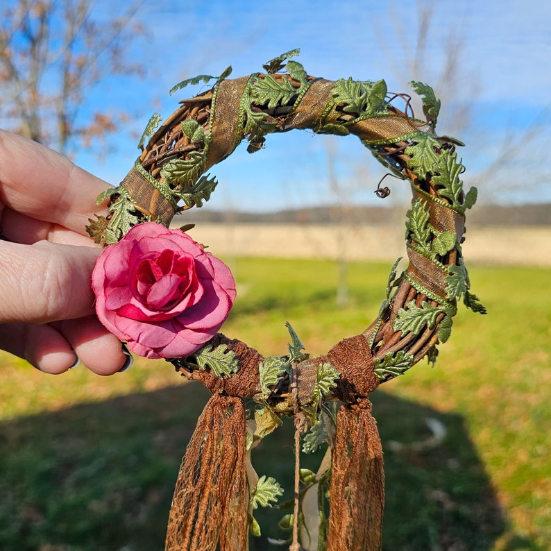 May include: A brown twig wreath with green leaves and a pink fabric rose. The wreath is decorated with brown ribbon and has long brown tassels hanging from the bottom.