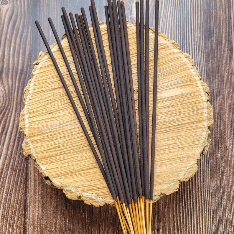May include: A bundle of dark brown incense sticks with light brown tips, arranged on a round, light brown wooden slab. The incense sticks are straight and evenly spaced, creating a visually appealing pattern. The background is a dark brown wooden surface.