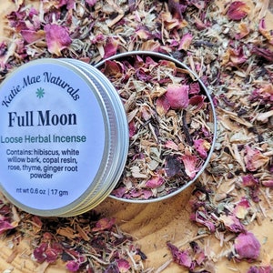May include: A silver tin with a white label that reads "Katie Mae Naturals Full Moon Loose Herbal Incense" and lists the ingredients: hibiscus, white willow bark, copal resin, rose, thyme, ginger root powder. The tin is open and filled with a blend of dried herbs and rose petals. The net weight is 0.6 oz or 17 gm.