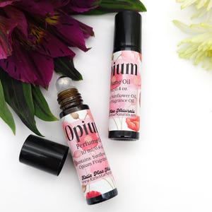 Puede incluir: Dos botellas de aceite de perfume Opium de 10 ml con un dise&ntilde;o floral y el texto "Opium Perfume Oil 10 ml/0.4 oz Contains: Sunflower Oil, Opium Fragrance Oil Katie Mae Naturals www.KatieMaeNaturals.net"
