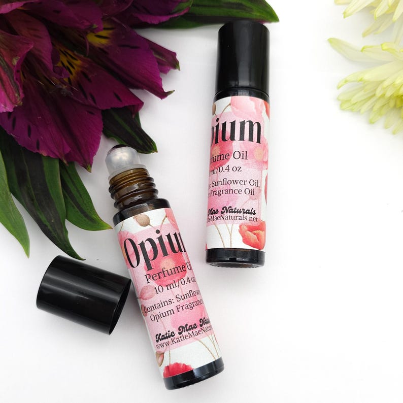 Puede incluir: Dos botellas de aceite de perfume Opium de 10 ml con un dise&ntilde;o floral y el texto "Opium Perfume Oil 10 ml/0.4 oz Contains: Sunflower Oil, Opium Fragrance Oil Katie Mae Naturals www.KatieMaeNaturals.net"