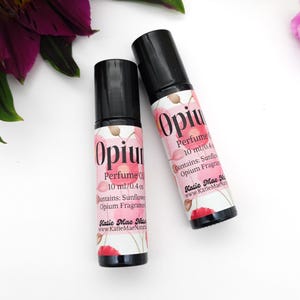 Puede incluir: Dos peque&ntilde;os frascos de vidrio con tapas de rodillo negras y etiquetas que dicen "Opium Perfume Oil 10 ml/0.4 oz Contains: Sunflower Opium Fragrance Katie Mae Naturals www.KatieMaeNaturals.com". Las etiquetas tienen un dise&ntilde;o floral rosa.