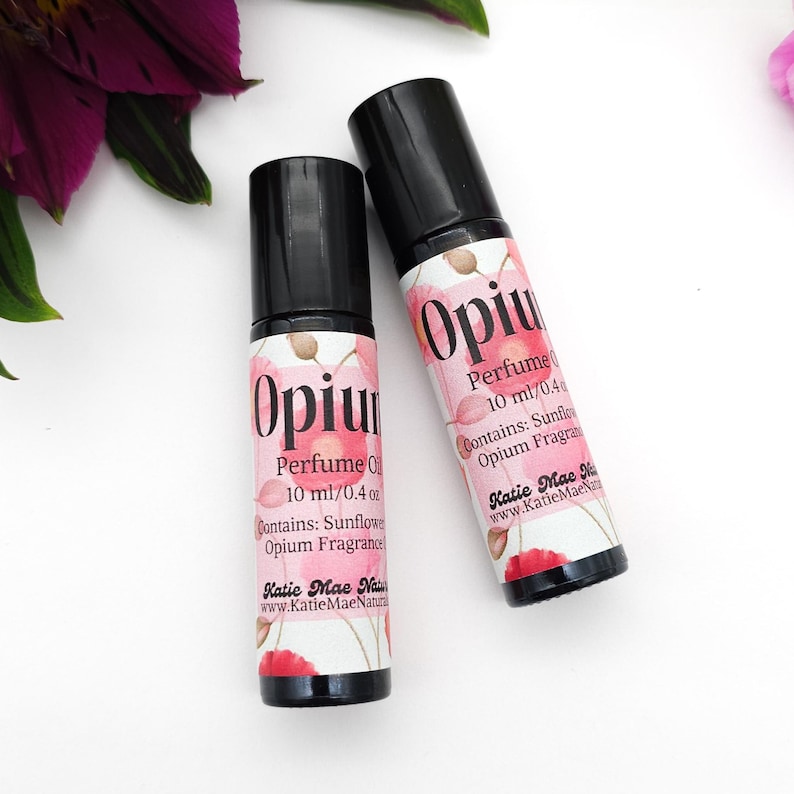 Puede incluir: Dos peque&ntilde;os frascos de vidrio con tapas de rodillo negras y etiquetas que dicen "Opium Perfume Oil 10 ml/0.4 oz Contains: Sunflower Opium Fragrance Katie Mae Naturals www.KatieMaeNaturals.com". Las etiquetas tienen un dise&ntilde;o floral rosa.