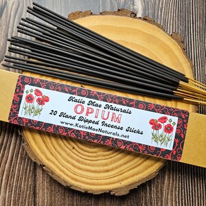 May include: A package of 20 hand-dipped incense sticks labeled "Opium" by Katie Mae Naturals. The sticks are black with gold tips, presented on a wooden slice. The packaging features red poppy illustrations and decorative patterns.