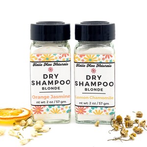 May include: Two small glass jars with white powder inside. The jars are labeled "Katie Mae Naturals Dry Shampoo Blonde" with the scents "Orange Jasmine" and "Lemon Chamomile". Each jar contains 2 ounces or 57 grams of dry shampoo.