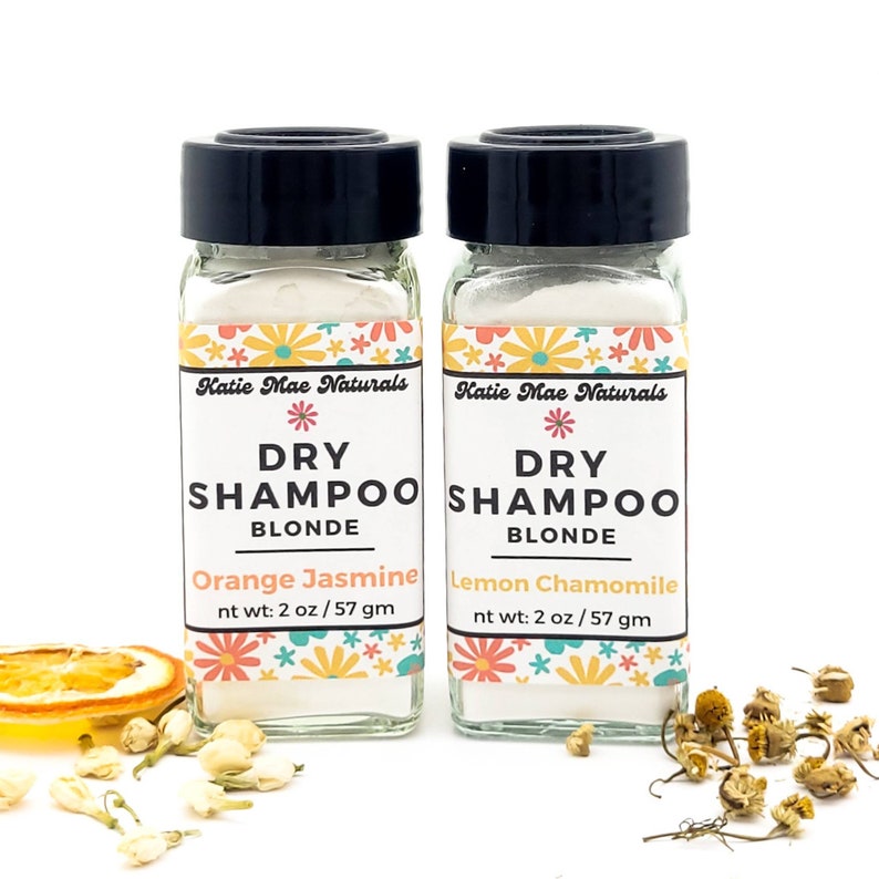 May include: Two small glass jars with white powder inside. The jars are labeled "Katie Mae Naturals Dry Shampoo Blonde" with the scents "Orange Jasmine" and "Lemon Chamomile". Each jar contains 2 ounces or 57 grams of dry shampoo.