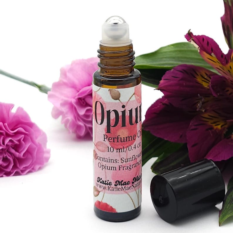 Puede incluir: Un frasco de vidrio marr&oacute;n con una bola de rodillo plateada. La etiqueta es blanca con flores de amapola rosadas y texto negro que dice "Opium Perfume Oil 10 ml/0.4 oz Contains: Sunflower Opium Fragrance Katie Mae Naturals www.KatieMaeNaturals.com".