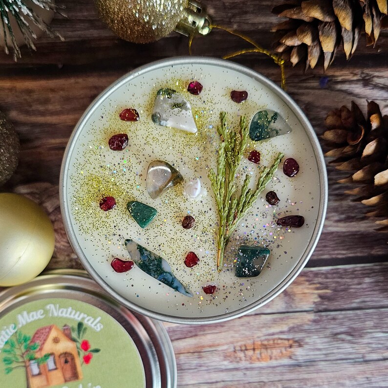 May include: A white candle with green and red glitter and small green, white, and red stones embedded in the wax. A sprig of green foliage is embedded in the center of the candle.