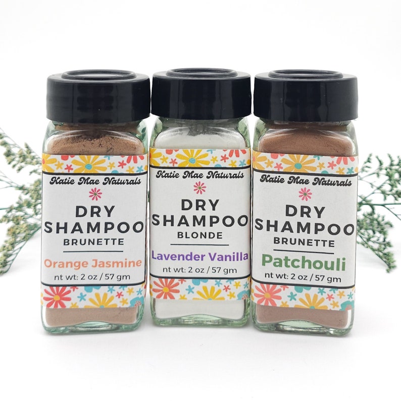 May include: Three small glass jars with black lids and colorful labels. Each jar contains a different color dry shampoo powder. The labels read "Dry Shampoo" with a hair color and scent listed below. The scents are "Orange Jasmine", "Lavender Vanilla", and "Patchouli".