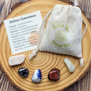 May include: A set of six gemstones in a small wooden bowl, with a white cloth bag with the text "Katie Mae Naturals" printed on it. The gemstones are feldspar, hematite, sodalite, red tiger's eye, and aquamarine. A card with the text "Mabon Gemstones" and a list of the gemstones and their properties is also included.