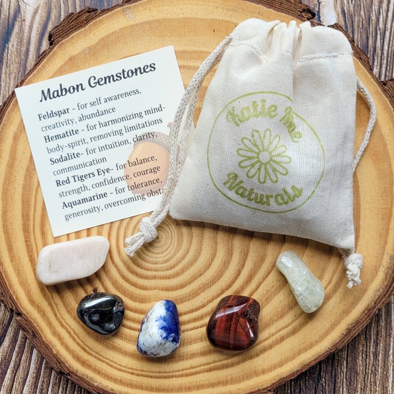 May include: A set of six gemstones in a small wooden bowl, with a white cloth bag with the text "Katie Mae Naturals" printed on it. The gemstones are feldspar, hematite, sodalite, red tiger's eye, and aquamarine. A card with the text "Mabon Gemstones" and a list of the gemstones and their properties is also included.