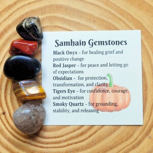 May include: A set of six gemstones, including black onyx, red jasper, obsidian, tiger's eye, and smoky quartz, arranged on a wooden surface. The gemstones are accompanied by a card with the text "Samhain Gemstones" and descriptions of each stone's properties.