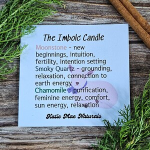 May include: A blue and white card with the text "The Imbolc Candle" and a list of crystals and herbs used in the candle, including Moonstone, Smoky Quartz, and Chamomile. The text "Katie Mae Naturals" is at the bottom of the card.