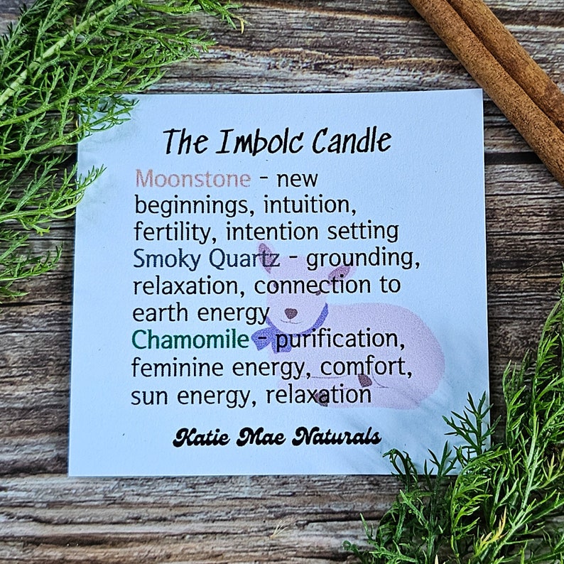 May include: A blue and white card with the text "The Imbolc Candle" and a list of crystals and herbs used in the candle, including Moonstone, Smoky Quartz, and Chamomile. The text "Katie Mae Naturals" is at the bottom of the card.