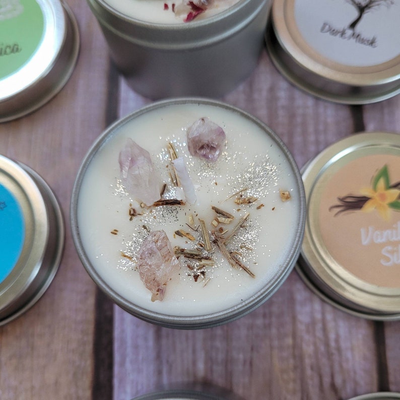 Puede incluir: Una vela blanca con una tapa plateada y una etiqueta blanca. La vela est&aacute; decorada con peque&ntilde;os cristales rosas y transparentes y purpurina plateada. La vela est&aacute; en un recipiente de hojalata.