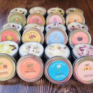 Puede incluir: Surtido de peque&ntilde;as latas redondas de metal con tapas. Cada lata contiene una vela de cera de soja con varios aromas y elementos decorativos. Las etiquetas de las latas presentan una variedad de dise&ntilde;os y textos, incluyendo "Katie Mae Naturals", "Magic Potion", "Apples & Oak", "Angelica" y "Orange Clove".