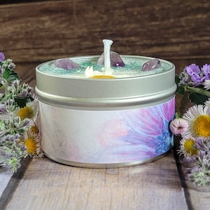 May include: A silver tin candle with a floral design label. The candle's surface is decorated with purple stones, green glitter, and a white wick. The candle is surrounded by flowers with purple and yellow blooms.