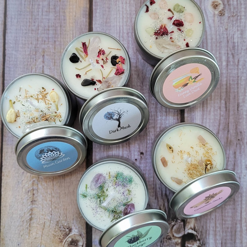 Puede incluir: Surtido de nueve peque&ntilde;as latas redondas de metal con tapas. Cada lata contiene una vela blanca con varios cristales, hierbas y purpurina. Las tapas tienen etiquetas con diferentes aromas, incluyendo "Moon Garden", "Dark Musk", "Honey Vanilla Love Dose", "Cinnamon Sticks" y "Blackberry Sage".