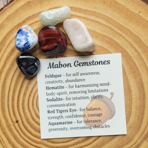 May include: A card with the text "Mabon Gemstones" and a list of gemstones and their properties. The card is surrounded by six small gemstones in various colors, including blue, green, red, black, and white.