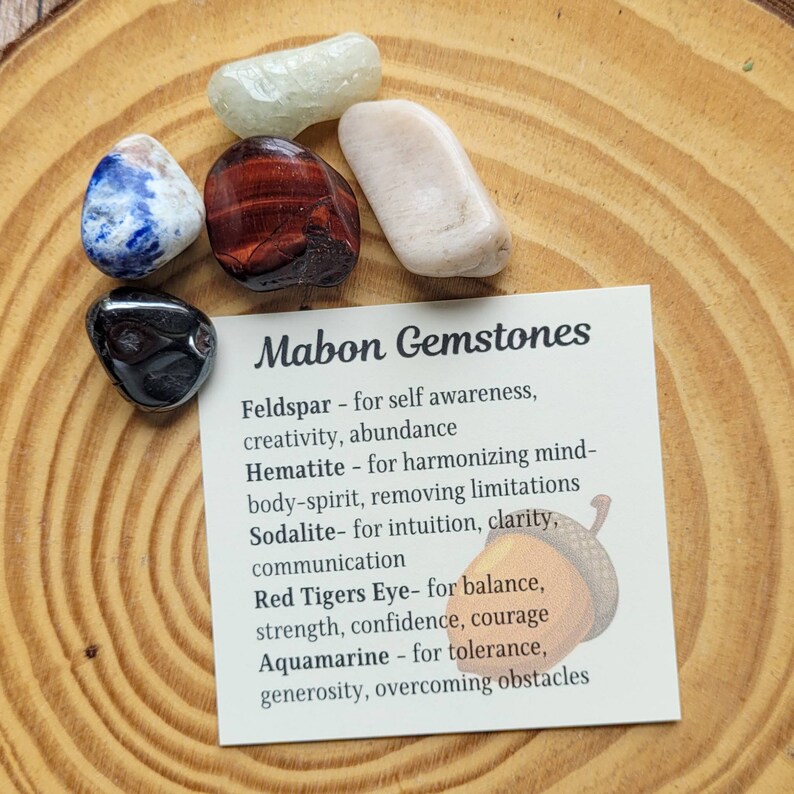 May include: A card with the text "Mabon Gemstones" and a list of gemstones and their properties. The card is surrounded by six small gemstones in various colors, including blue, green, red, black, and white.