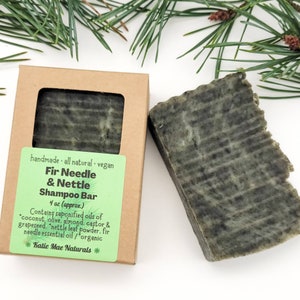 May include: A brown cardboard box with a green label that reads "handmade - all natural - vegan Fir Needle & Nettle Shampoo Bar 4 oz (approx.) Contains saponified oils of "coconut, olive, almond, castor & grapeseed. "nettle leaf powder, fir needle essential oil/organic Katie Mae Naturals *"  A green bar of soap is sitting next to the box.
