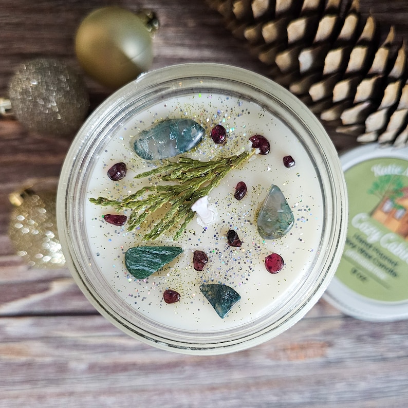 Puede incluir: Una vela de cera de soja vertida a mano en un tarro de vidrio, decorada con piedras preciosas verdes y azules, cuentas rojas y una ramita de vegetaci&oacute;n. La vela est&aacute; etiquetada como "Cozy Cabin". El brillo a&ntilde;ade brillo.