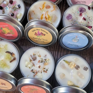 Puede incluir: Surtido de peque&ntilde;as velas redondas de cera de soja hechas a mano en latas de metal. Cada vela tiene un aroma natural diferente y elementos decorativos, como flores secas, cristales y purpurina. Las etiquetas de las latas muestran el nombre de la marca "Katie Mae Naturals" y los nombres de los aromas, entre ellos "Butterfly Flower", "Magic Potion" y "Yuletide".